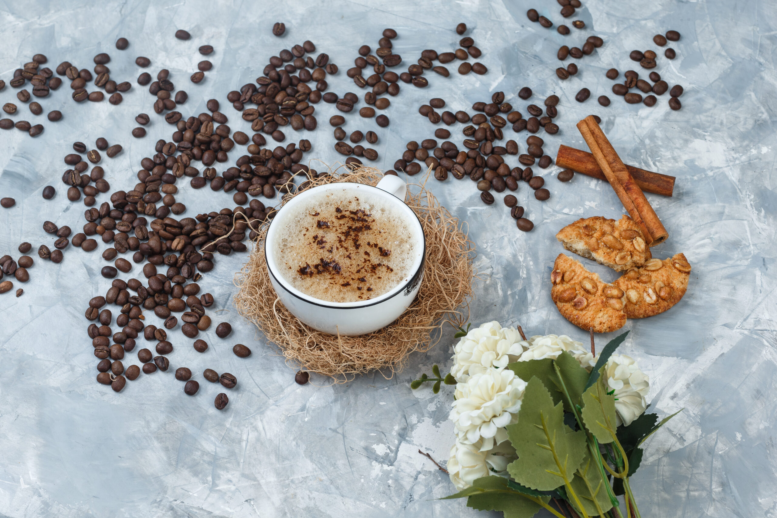 high angle view coffee in cup with cookies, coffee beans, flowers, cinnamon sticks on grey plaster background